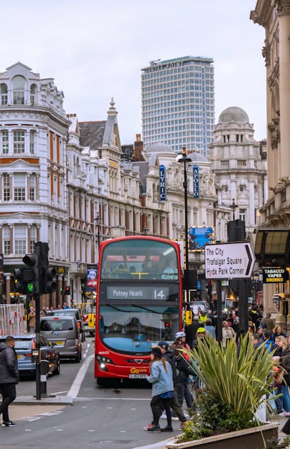 In the foreground, a group of pedestrians, including a woman wearing a blue jacket and a man in a darker coat, cross a busy city street paved with asphalt. To the right, a large green plant with long,
