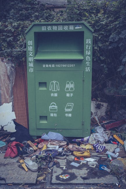 A collection of three large plastic waste bags positioned on the pavement near a black metal fence, with one bag leaning against the fence and the others lying flat on the ground. The bags appear to b